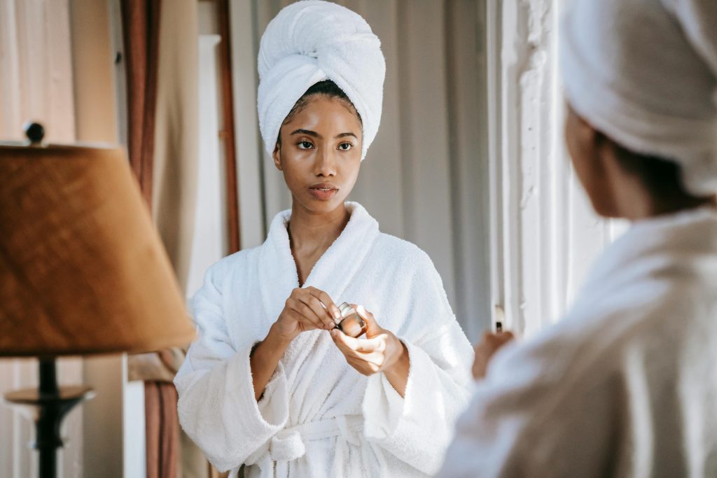 Programmes Crop feminine ethnic female in soft robe with cosmetic product looking in mirror in house room