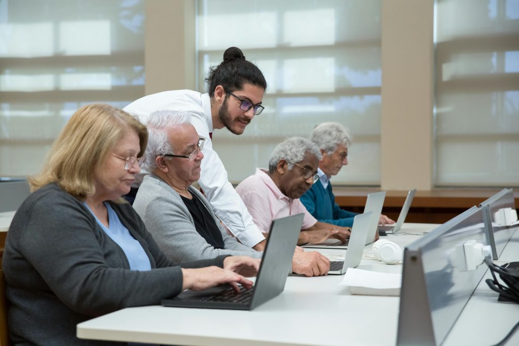 Programmes Senior adults use laptops in a classroom setting, guided by a young instructor.