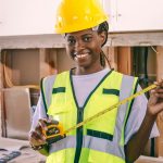 Smiling female engineer holding a measuring tape, wearing PPE and hardhat indoors.